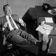 Black and white photo of man laughing with feet up on desk, drinking glass of milk